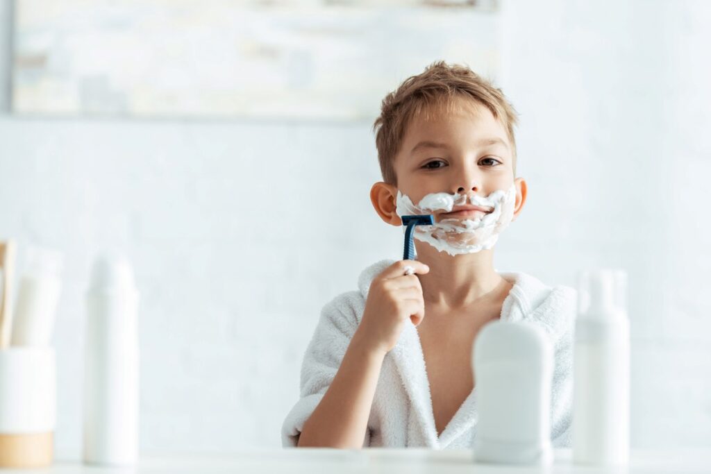 Αγόρι μικρής ηλικίας στο μπάνιο με αφρό στο πρόσωπο που μιμείται το ξύρισμα μπροστά στον καθρέφτη, συμβολίζοντας πρώιμα σημάδια ανάπτυξης και πρώιμη αδρεναρχή.